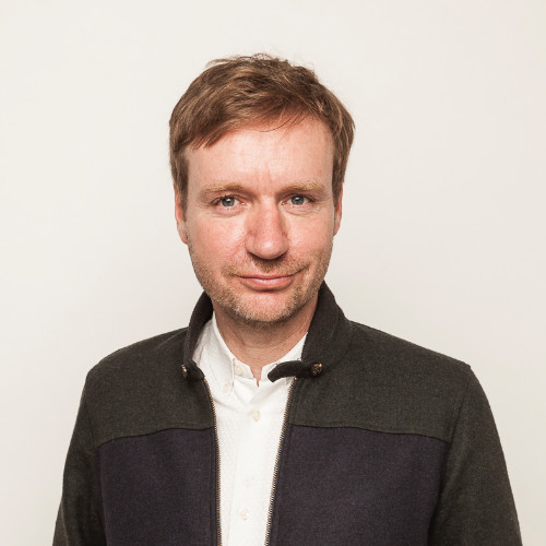 Porträt eines lächelnden Mannes mit hellem Hintergrund, der eine weiße Bluse und eine dunkle Jacke trägt.