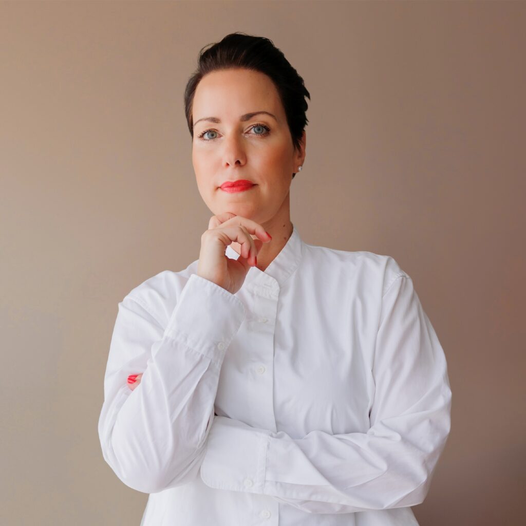 Frau in weißem Hemd vor neutralem Hintergrund, nachdenklich mit Hand am Kinn.