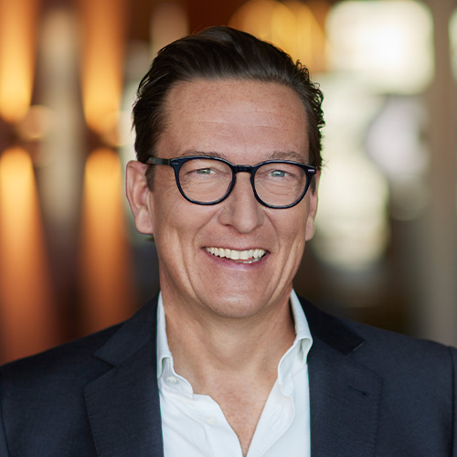 Eine lächelnde Person mit Brille in einem Anzug, unscharfer Hintergrund, Innenaufnahme.