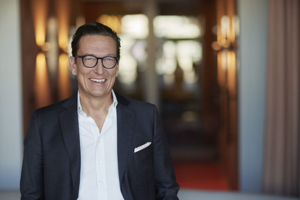 Geschäftsmann in Anzug und Brille lächelt in die Kamera, Hintergrund unscharf.