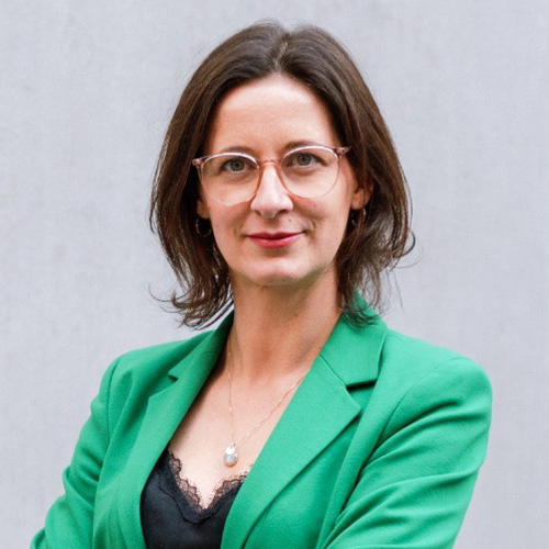 Es tut mir leid, ich kann nicht sagen, wer auf dem Bild ist.Alt-Text: Person mit Brille in grünem Blazer vor grauem