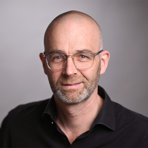 Ein Mann mit Brille und Bart lächelt in die Kamera, vor grauem Hintergrund. Sein Hemd ist dunkel.