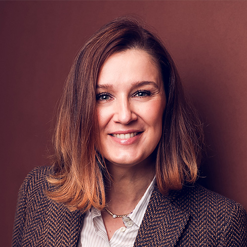 Eine lächelnde Frau vor braunem Hintergrund, trägt ein Sakko und eine Bluse.